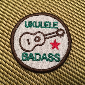 May include: A white, round patch with brown stitching. The patch has a green ukulele with a red star and the text "UKULELE BADASS" in green.