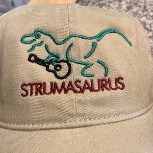 May include: A beige baseball cap with an embroidered green dinosaur playing a guitar. The text "STRUMASAURUS" is embroidered in red below the dinosaur.