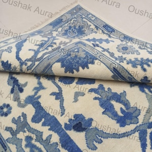 Pode incluir: Um tapete dobrado, branco sujo, com um padrão floral azul detalhado. O tapete apresenta uma borda com um design repetitivo e uma franja ao longo de uma borda. O design inclui flores e folhas estilizadas.