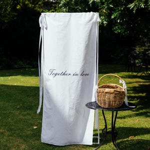 May include: A tall white fabric banner featuring the black text Together in love, suspended by white ribbons. Beside it sits a round black metal table holding a woven wicker basket with a handle, decorated with a white ribbon tied to the side.