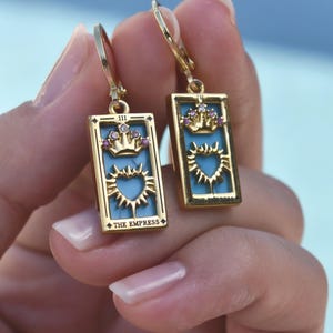 May include: Gold-tone earrings featuring a rectangular design with a light blue background. Each earring has a crown and heart design with the text "THE EMPRESS". The earrings are held up by a gold-colored lever back.