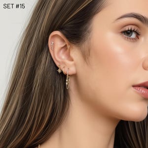 May include: A collection of gold earrings, including a star-shaped stud, a hoop with a chain, and a series of small studs. The earrings are worn on the ear of a person with long, light brown hair. The text "SET #15" is in the upper left corner.