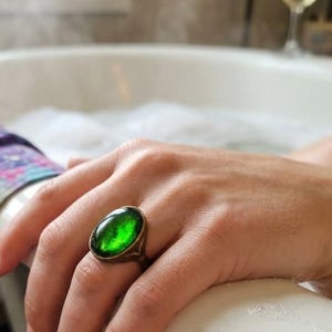 May include: A ring featuring a large, oval-shaped, vibrant green cabochon stone set in a metallic band. The stone has a smooth, polished surface with deep emerald tones and subtle internal variations, securely held by a simple metal setting.