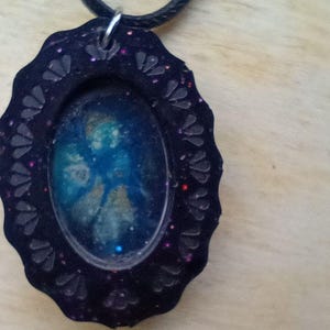 May include: A black oval pendant necklace with a decorative frame. The pendant features a blue and gold abstract design within a clear resin. The frame is embellished with glitter and a scalloped edge. The necklace cord is black.