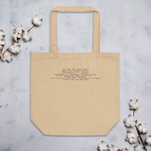 Puede incluir: Bolso tote beige con un fragmento de código impreso en negro. El bolso tiene forma rectangular con una abertura ancha y dos asas. Ramas de algodón con flores blancas en el fondo.