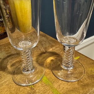 May include: Two clear glass wine glasses with a spiral stem design. The glasses sit on a wooden surface. The background includes a dark blue wall and a glimpse of a yellow candle.