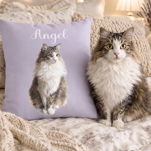 May include: A lilac pillow with a cat portrait and the name "Angel" in white script. A fluffy, long-haired cat with brown, white, and gray fur sits beside the pillow. The scene is set on a floral patterned bedspread.