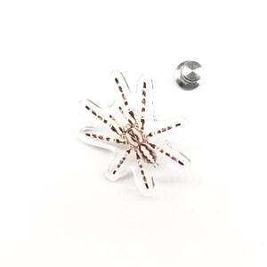 Pode incluir: Um alfinete transparente em forma de aranha com uma ilustração detalhada de uma aranha em tons de castanho e branco. O alfinete tem um suporte prateado para fixação. O design está em um fundo branco.