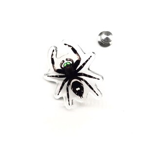 Pode incluir: Um alfinete de acrílico transparente com uma aranha preta e branca com olhos verdes. O alfinete tem um verso prateado. As pernas da aranha estão estendidas. O design é detalhado.