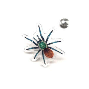 Pode incluir: Um broche transparente em forma de aranha com um design bordado detalhado. A aranha apresenta uma cabeça verde, um abdómen castanho e pernas azuis. Uma parte traseira prateada é visível.