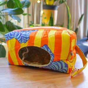May include: A colorful, quilted tissue box cover with a vibrant design. The cover features vertical orange and yellow stripes, with blue and white wave-like patterns. It has a zippered top and a small handle with a gold-colored clasp.