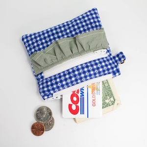 May include: A handmade blue and white gingham fabric wallet with a green ruffle detail. The wallet has two zippered compartments, one holding a Costco membership card and a dollar bill. Loose change is visible.
