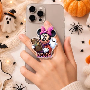 May include: A phone grip featuring a cartoon mouse character wearing glasses and a purple bow, holding a book with a small ghost beside it. The name BELLA is printed in orange and pink letters at the bottom of the acrylic design on a gray phone.