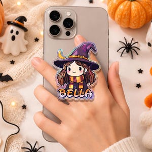 May include: A phone grip featuring a cartoon illustration of a character wearing a purple witch hat and a striped scarf. The character has brown hair and a smiling expression. The name BELLA is printed in bold orange letters at the bottom.