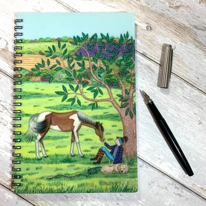 May include: A spiral bound notebook featuring a colourful illustration of a girl stroking a horse in a field. The horse is brown and white with a black mane and tail. The girl is wearing a blue shirt and a red hat. The background is a green field with a tree and a purple flower patch.