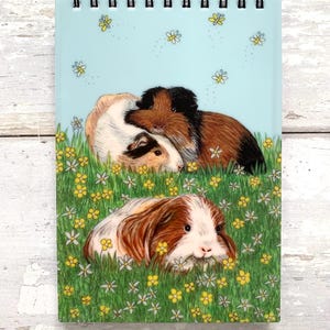 May include: A spiral-bound notepad featuring an illustration of guinea pigs in a field of grass and flowers. The guinea pigs are in shades of brown, white, and black. The background is a light blue with yellow butterflies.