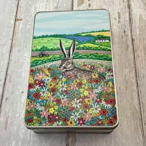 May include: A rectangular tin featuring a detailed illustration of a hare amidst a vibrant wildflower meadow. The background shows a rural scene with fields, a tractor, and a blue sky. The tin is likely for storage.