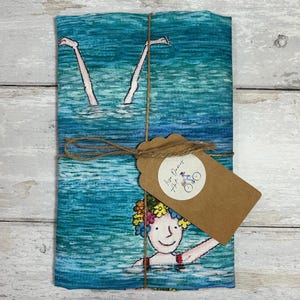 May include: Folded blue and teal fabric featuring a swimming illustration with legs in the water. A brown tag is tied with twine. The tag has a circular logo with text and a bicycle. The fabric is tied with twine.