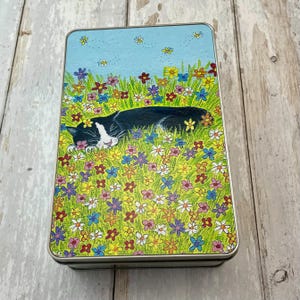 May include: A rectangular metal tin with a painted design. The lid features a black and white cat sleeping in a field of colourful flowers. The background is a light blue sky with small yellow butterflies.