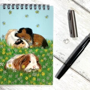 May include: A spiral-bound notepad with a light blue cover featuring an illustration of guinea pigs in a field of flowers. A black fountain pen and a silver pen cap are next to the notepad on a white wooden surface.