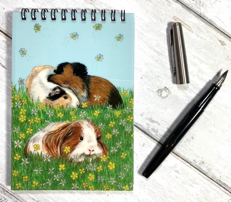 May include: A spiral-bound notepad with a light blue cover featuring an illustration of guinea pigs in a field of flowers. A black fountain pen and a silver pen cap are next to the notepad on a white wooden surface.