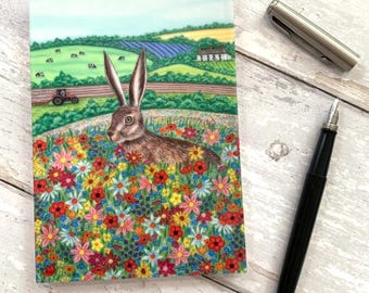 May include: A spiral-bound notepad with a colorful illustration of a hare in a field of wildflowers. A black fountain pen and a silver pen are next to the notepad on a white wooden surface. The notepad features a rural landscape.