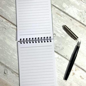 May include: An open, spiral-bound notepad with lined white pages. A black pen and a silver pen cap are next to the notepad on a light wood-grain surface. The notepad is open to reveal the lined pages.