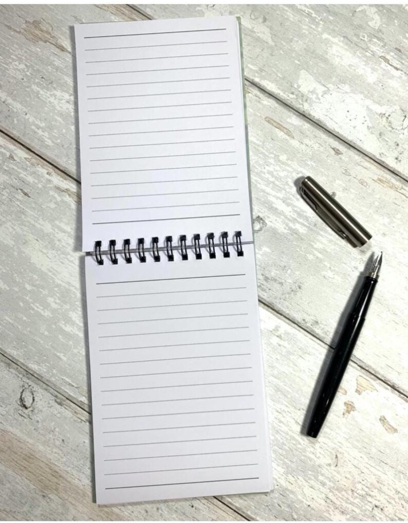 May include: An open, spiral-bound notepad with lined white pages. A black pen and a silver pen cap are next to the notepad on a light wood-grain surface. The notepad is open to reveal the lined pages.