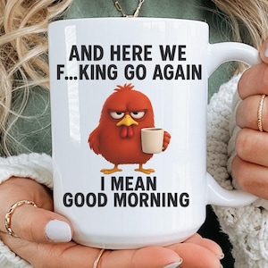 May include: A white ceramic mug featuring a graphic of a grumpy red chicken holding a small cup. The text above the chicken reads AND HERE WE F...KING GO AGAIN and the text below reads I MEAN GOOD MORNING. The mug has a curved handle and a smooth finish.