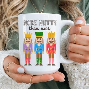 May include: White ceramic mug with the text "MORE NUTTY than nice" and illustrations of three toy nutcracker figures in purple, blue, and red uniforms. The mug has a large handle.