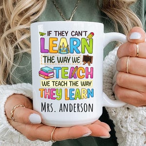 Könnte beinhalten: Weißer Keramikbecher mit großem Henkel, mit farbigem Text, der lautet: "IF THEY CAN'T LEARN THE WAY WE TEACH WE TEACH THE WAY THEY LEARN MRS. ANDERSON." Das Design umfasst schulische Grafiken wie Bleistift, Bücher und eine Tafel.