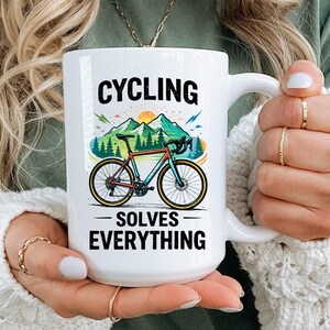 以下が含まれることがあります： 「CYCLING SOLVES EVERYTHING」の文字が黒で書かれた白いセラミックマグ。マグには、自転車と山並み、昇る太陽のカラフルなグラフィックが描かれています。マグは人が持っています。