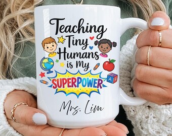 May include: White ceramic mug with the text "Teaching Tiny Humans is My SUPERPOWER" in black script and colourful graphics. The mug features illustrations of children, an apple, a globe, and other school-related items. The name "Mrs. Lim" is printed below.