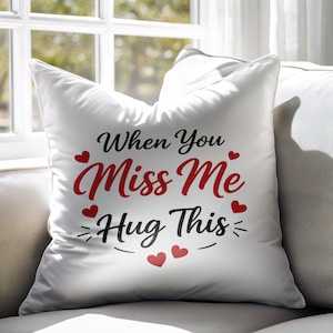 Puede incluir: Cojín decorativo blanco con el texto "When You Miss Me Hug This" en negro y rojo. El cojín presenta detalles de corazones rojos y está colocado en un sofá gris cerca de una ventana.
