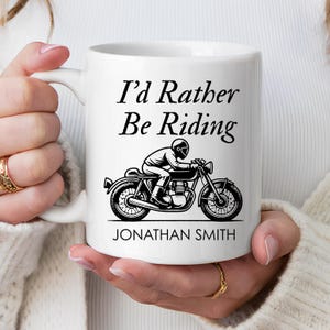 以下が含まれることがあります： 「I'd Rather Be Riding」の文字と、バイクとライダーの黒いイラストが描かれた白いセラミックマグカップ。「JONATHAN SMITH」という名前が下に印刷されています。マグカップにはカーブしたハンドルが付いています。