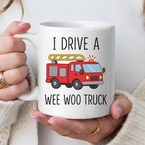 May include: White ceramic mug with the text "I DRIVE A WEE WOO TRUCK" and a cartoon illustration of a red fire truck. The fire truck has a yellow ladder and flashing lights. The mug is held by a person.
