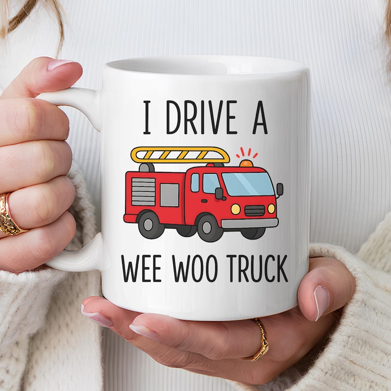 May include: White ceramic mug with the text "I DRIVE A WEE WOO TRUCK" and a cartoon illustration of a red fire truck. The fire truck has a yellow ladder and flashing lights. The mug is held by a person.