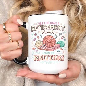 Op de afbeelding: Witte keramische mok met een zwarte handgreep en rand, met de tekst "YES I DO HAVE A RETIREMENT PLAN I PLAN ON KNITTING". Het ontwerp bevat illustraties van garen, breinaalden en knopen in pastelkleuren.