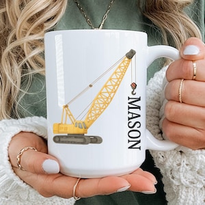 Könnte beinhalten: Weiße Keramiktasse mit einem gelben Kran-Design und dem Namen "MASON" vertikal in Schwarz gedruckt. Der Kran hat ein detailliertes Design mit einem roten Haken. Die Tasse hat einen großen Henkel und wird von einer Person gehalten.