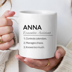 Op de afbeelding: Witte keramische mok met de naam "ANNA" in zwart, gevolgd door "Executive Assistant" en een lijst met taken. De mok wordt door een persoon vastgehouden.