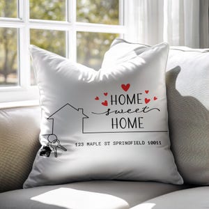 Könnte beinhalten: Weißes Zierkissen mit dem Schriftzug "HOME sweet HOME" in schwarzer Schrift und roten Herzen. Auf dem Kissen sind auch ein Hausumriss und Schlüssel abgebildet. Die Adresse "123 MAPLE ST SPRINGFIELD 10011" ist darunter aufgedruckt.