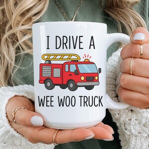 May include: White ceramic mug with the text "I DRIVE A WEE WOO TRUCK" and a red fire truck graphic. The fire truck has a yellow ladder and flashing lights. The mug is held by a person.