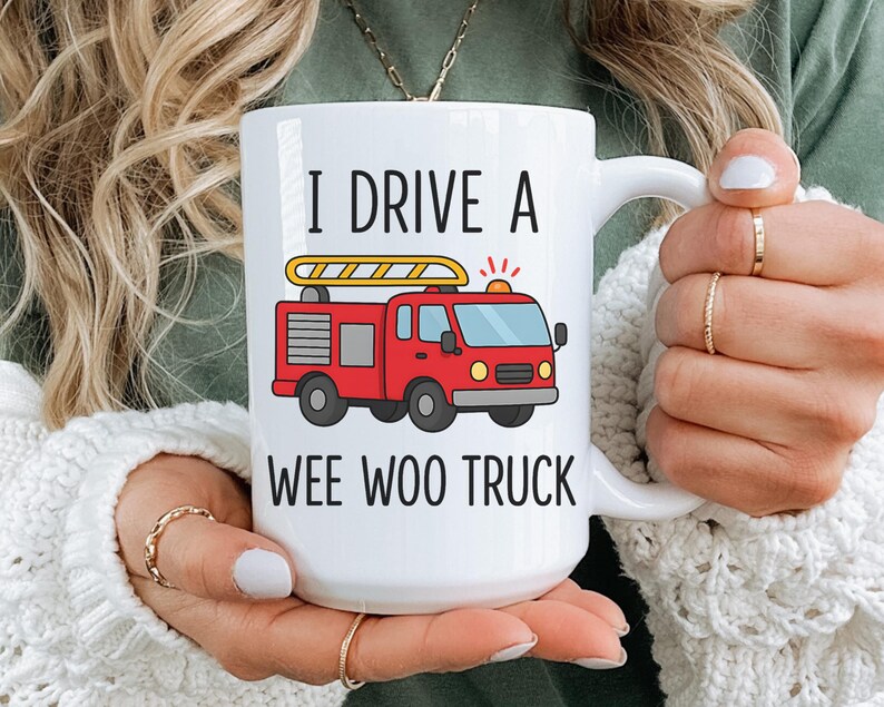 May include: White ceramic mug with the text "I DRIVE A WEE WOO TRUCK" and a red fire truck graphic. The fire truck has a yellow ladder and flashing lights. The mug is held by a person.