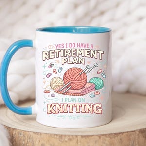 Op de afbeelding: Witte keramische mok met een blauwe handgreep en binnenkant. De mok heeft de tekst "YES I DO HAVE A RETIREMENT PLAN I PLAN ON KNITTING" met illustraties van garen, breinaalden en knopen. De mok staat op een houten oppervlak.