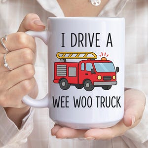 May include: White ceramic mug with the text "I DRIVE A WEE WOO TRUCK" and a cartoon illustration of a red fire truck with a yellow ladder. The mug is held by a person.