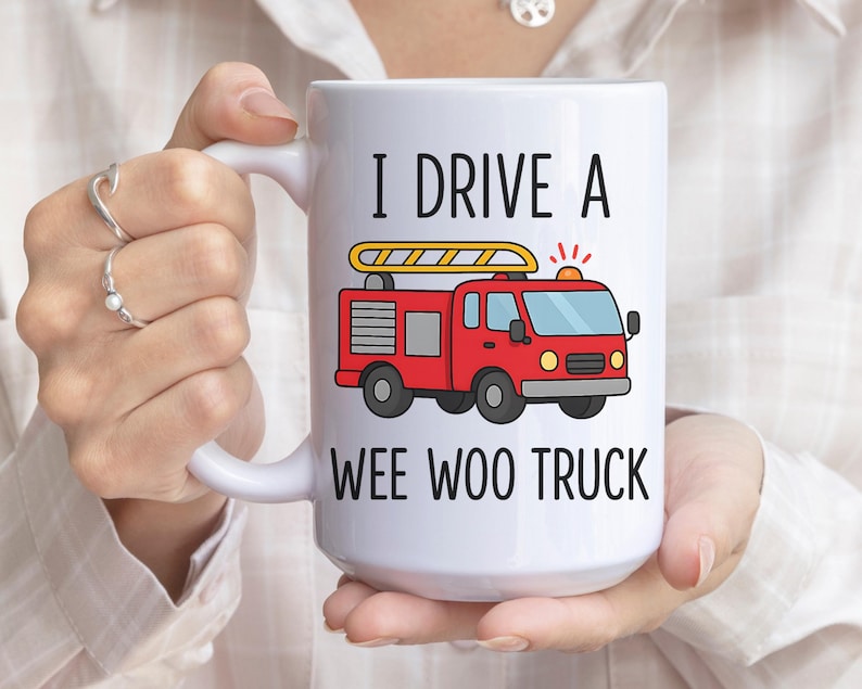 May include: White ceramic mug with the text "I DRIVE A WEE WOO TRUCK" and a cartoon illustration of a red fire truck with a yellow ladder. The mug is held by a person.