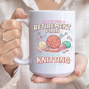 Op de afbeelding: Witte keramische mok met de tekst "YES I DO HAVE A RETIREMENT PLAN I PLAN ON KNITTING". De mok heeft illustraties van bollen wol, breinaalden en knopen. De mok wordt door een persoon vastgehouden.