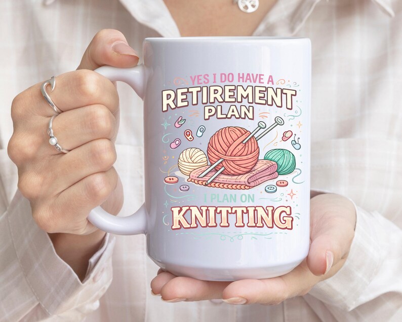 Op de afbeelding: Witte keramische mok met de tekst "YES I DO HAVE A RETIREMENT PLAN I PLAN ON KNITTING". De mok heeft illustraties van bollen wol, breinaalden en knopen. De mok wordt door een persoon vastgehouden.