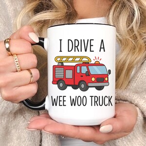 May include: White ceramic mug with a black handle and rim. The mug features the text "I DRIVE A WEE WOO TRUCK" and a cartoon illustration of a red fire truck with a yellow ladder. The mug is held by a person.