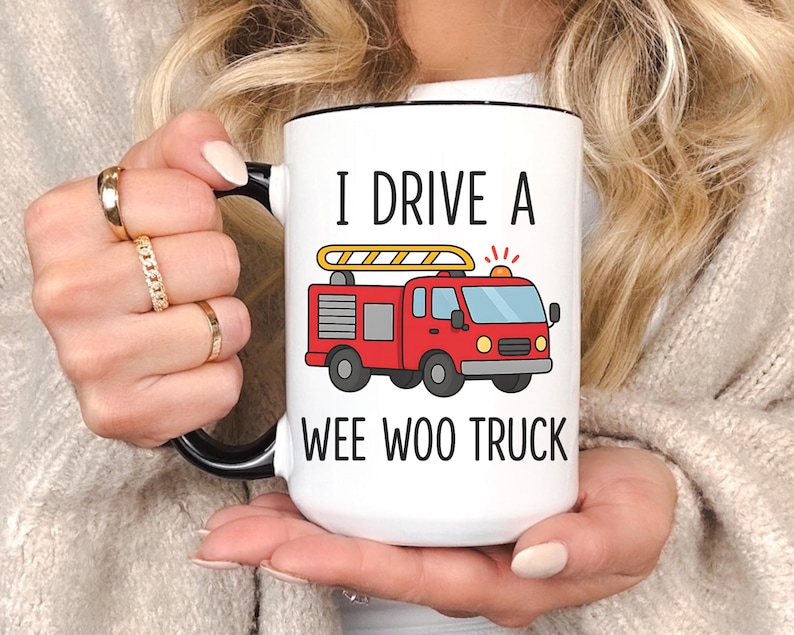 May include: White ceramic mug with a black handle and rim. The mug features the text "I DRIVE A WEE WOO TRUCK" and a cartoon illustration of a red fire truck with a yellow ladder. The mug is held by a person.